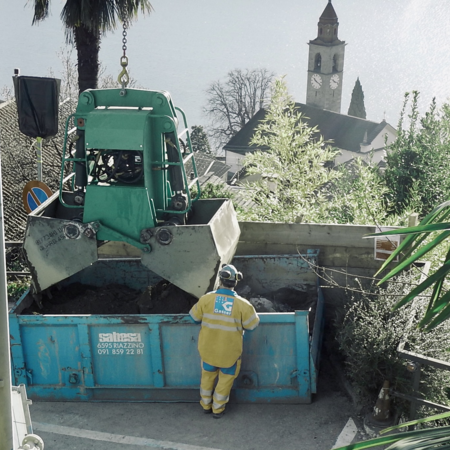 Diesel-hydraulischer Greifer, der in einem Garten in Hanglage Schutt transportiert. Im Hintergrund ist eine Kirche und das Meer zu sehen