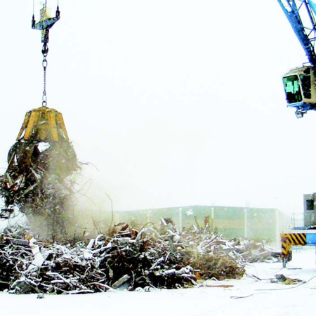 Diesel-hydraulic orange peel grab, remote-controlled type DHM on snow-covered terrain. It is attached to a blue crane and is transporting a large amount of scrap metal
